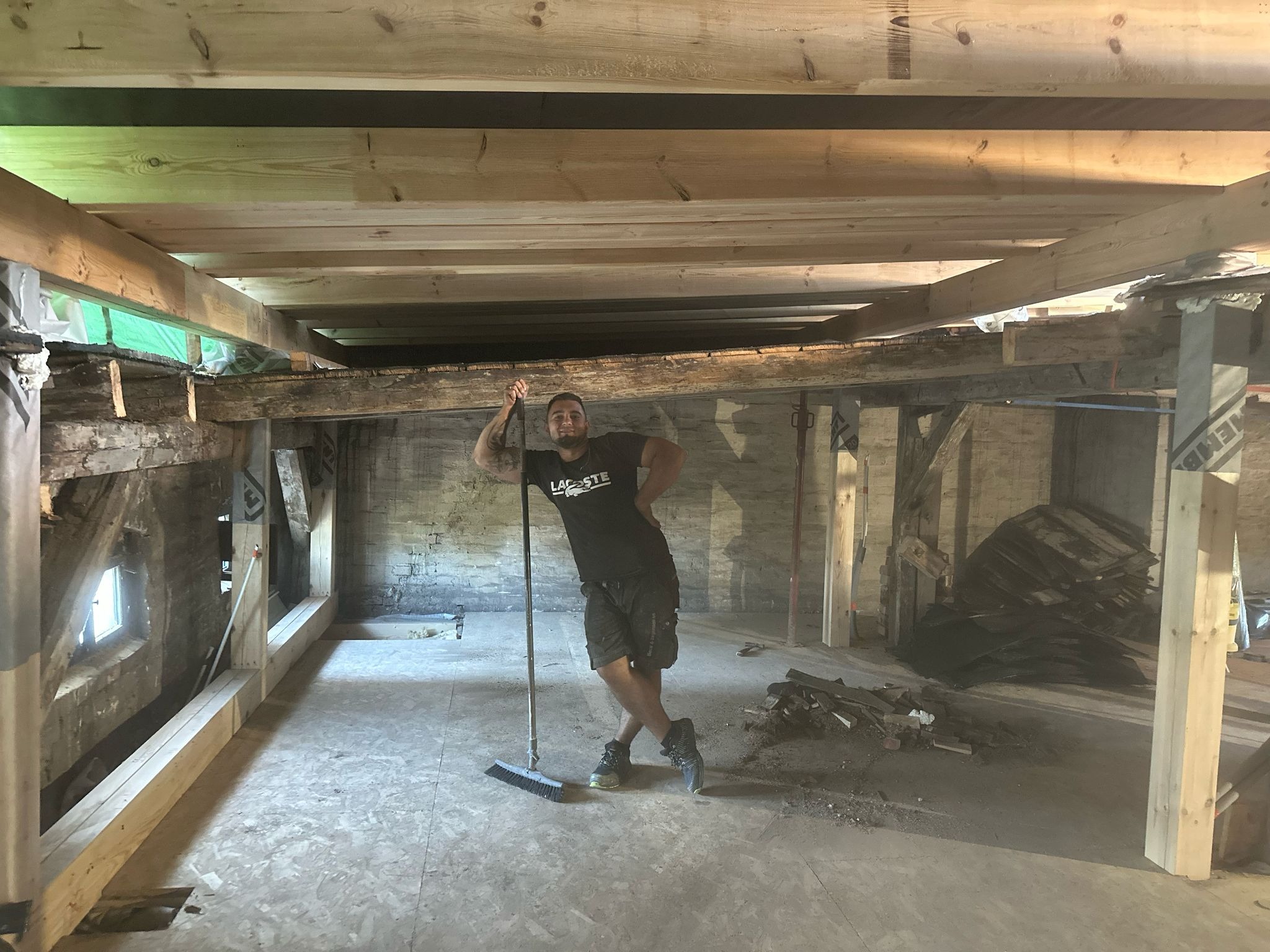 Uśmiechnięty mężczyzna oparty o miotłę w trakcie prac przygotowawczych do remontu poddasza, widoczne nowe drewniane belki stropowe i fragment starej ceglanej ściany.