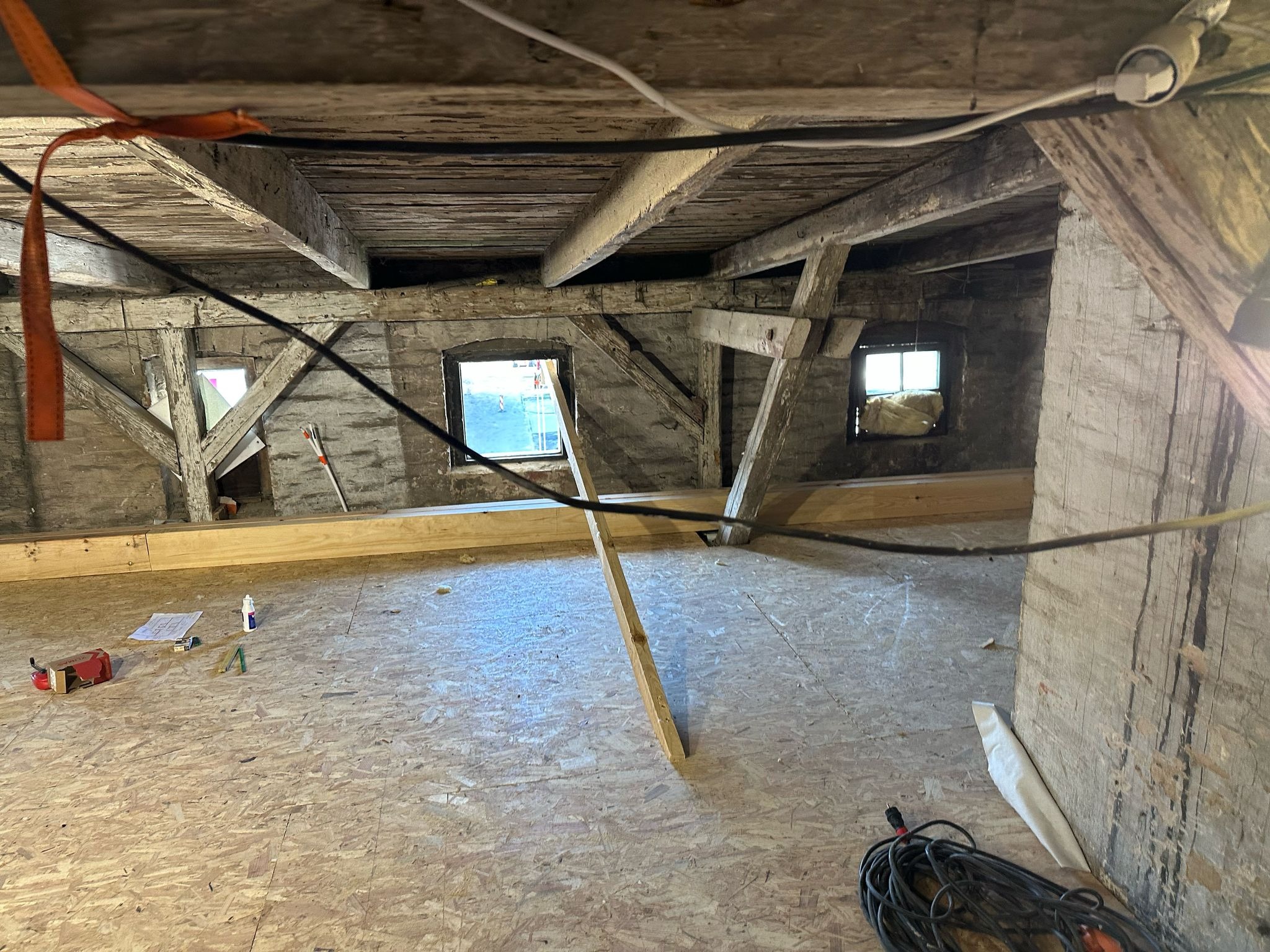 Poddasze w trakcie remontu: widoczne stare drewniane belki, fragment ściany z oknem, nowe panele podłogowe, narzędzia i materiały budowlane w nieładzie.