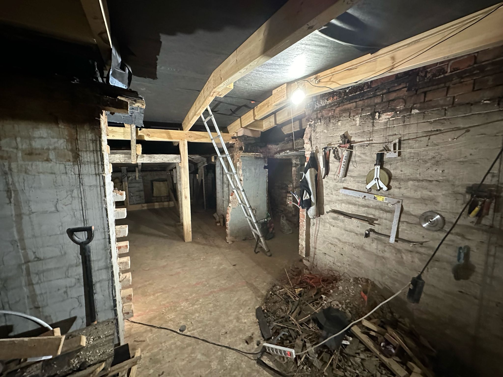 Wnętrze w trakcie przebudowy z odsłoniętą cegłą, nową drewnianą konstrukcją sufitu, drabiną opartą o ścianę i narzędziami zawieszonymi na ścianie.