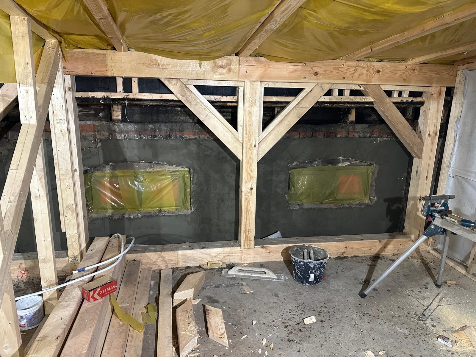 Wnętrze w trakcie budowy domu szkieletowego, widoczne drewniane elementy konstrukcyjne, zaizolowane okna folią ochronną i częściowo otynkowana ściana z cegieł, narzędzia budowlane i materiały...