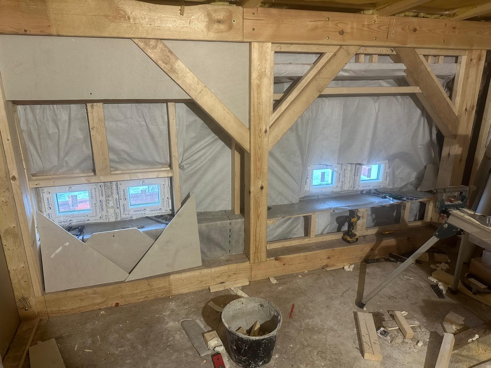 Wnętrze poddasza w trakcie adaptacji: widoczne drewniane elementy konstrukcyjne, ocieplenie folią, okna dachowe, przygotowane płyty gipsowe do montażu oraz narzędzia pracy.