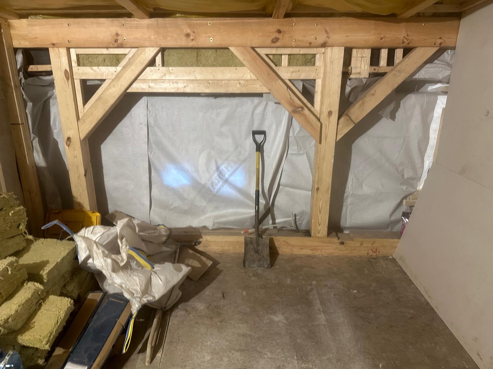 Wnętrze domu w trakcie budowy: drewniana konstrukcja szkieletowa z widoczną izolacją, szpachla i narzędzia, w tym łopata stojąca na podłodze.