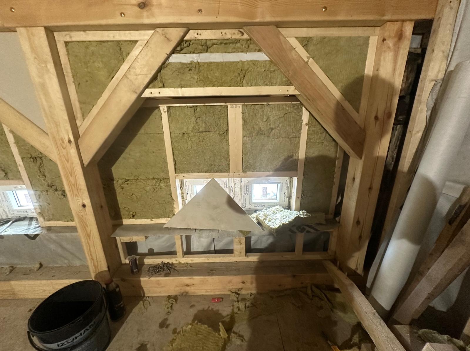 Poddasze w trakcie prac izolacyjnych: wełna mineralna między drewnianymi elementami konstrukcyjnymi, widoczne okno dachowe i fragmenty narzędzi oraz materiałów budowlanych. Uwagę zwraca trójkątny...