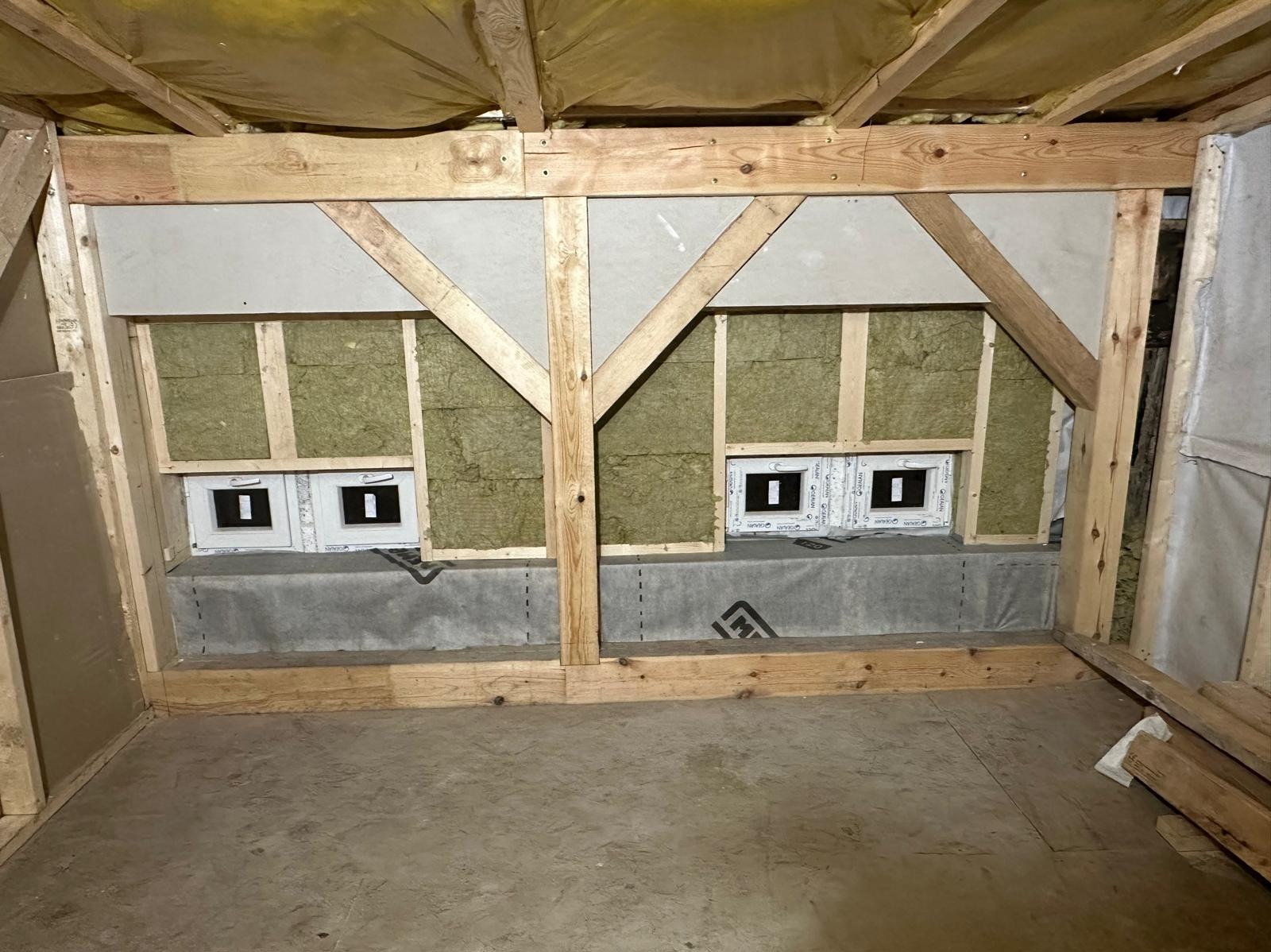 Wnętrze domu szkieletowego w trakcie budowy: widoczne drewniane ramy, ocieplenie z wełny mineralnej, zamontowane okna, betonowy fundament z hydroizolacją.