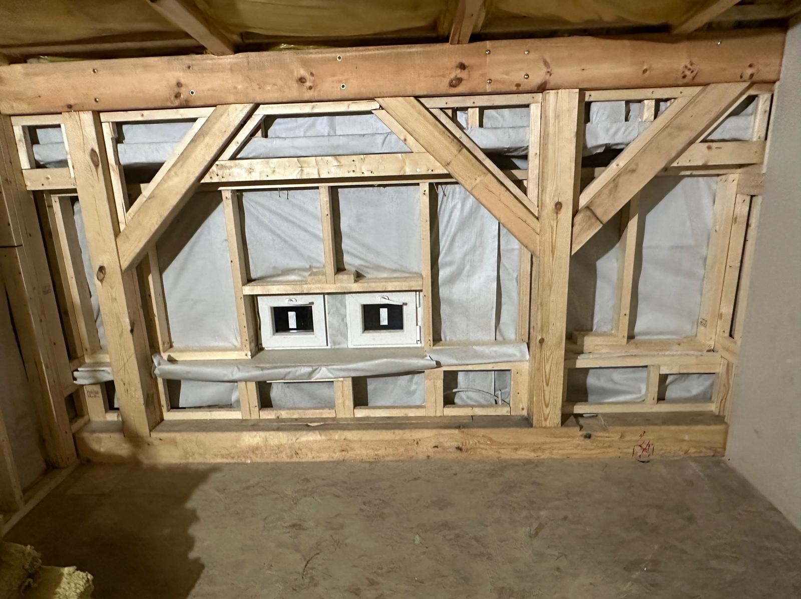 Wnętrze w trakcie budowy domu szkieletowego: widoczne drewniane ramy ścian z izolacją i wstawionymi oknami, surowa podłoga i fragment dachu.