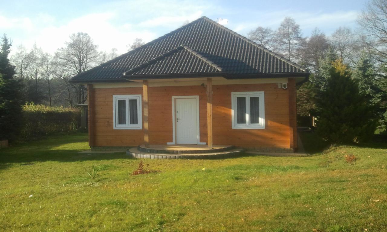 Parterowy dom z bali z ciemnym dachem, jasnymi oknami i drzwiami na tle zielonego trawnika i drzew. Widoczne detale wykończenia i naturalne oświetlenie.