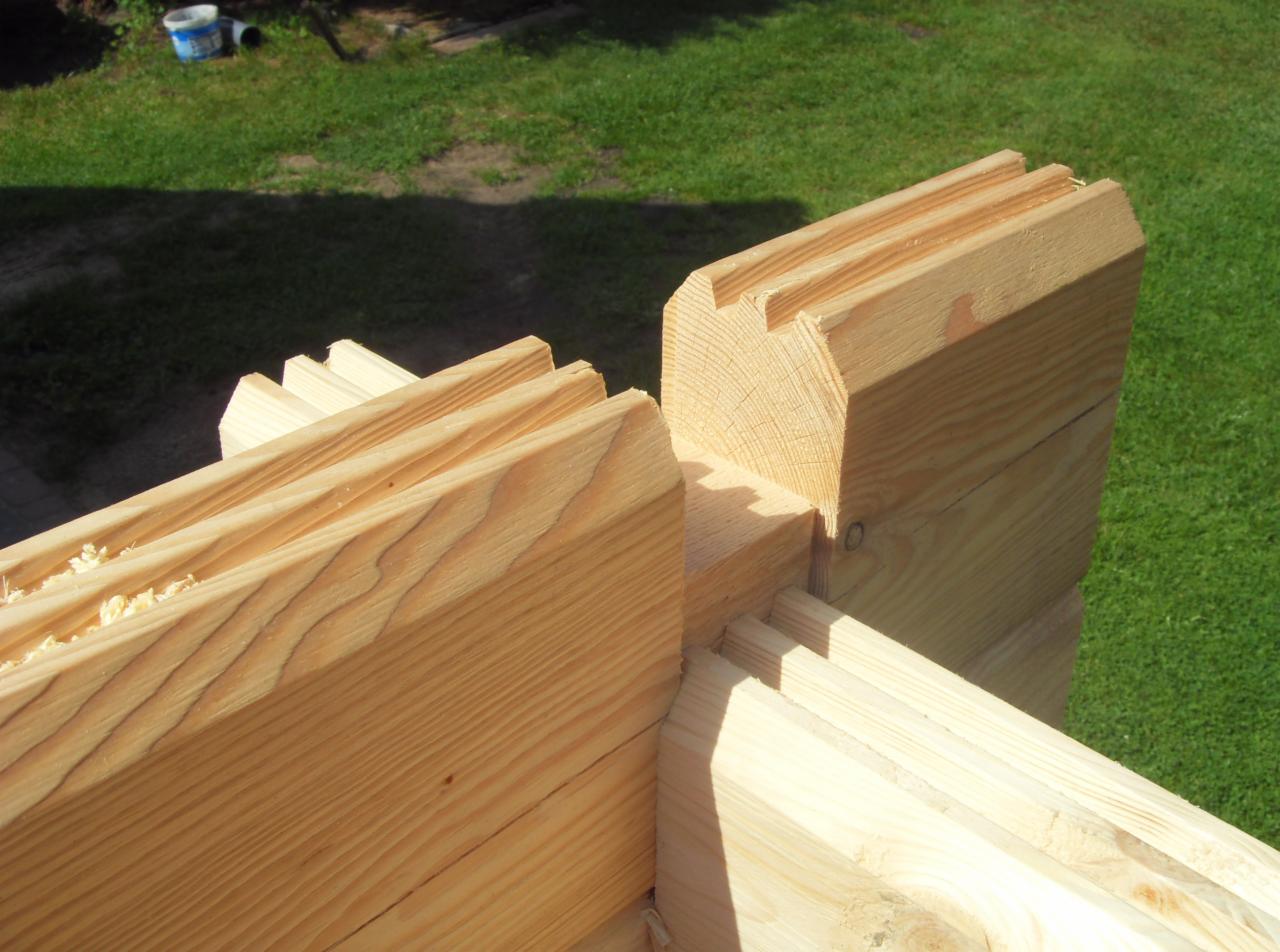 Detal drewnianej konstrukcji domu z bali, widoczne łączenie belek z charakterystycznym wyprofilowaniem. Tło stanowi zielona trawa.