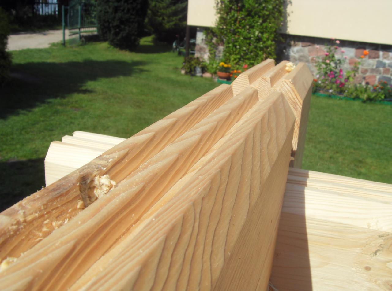 Profilowane deski z jasnego drewna, widoczne słoje i drobne wióry. Element konstrukcyjny na tle zielonego trawnika i budynku.
