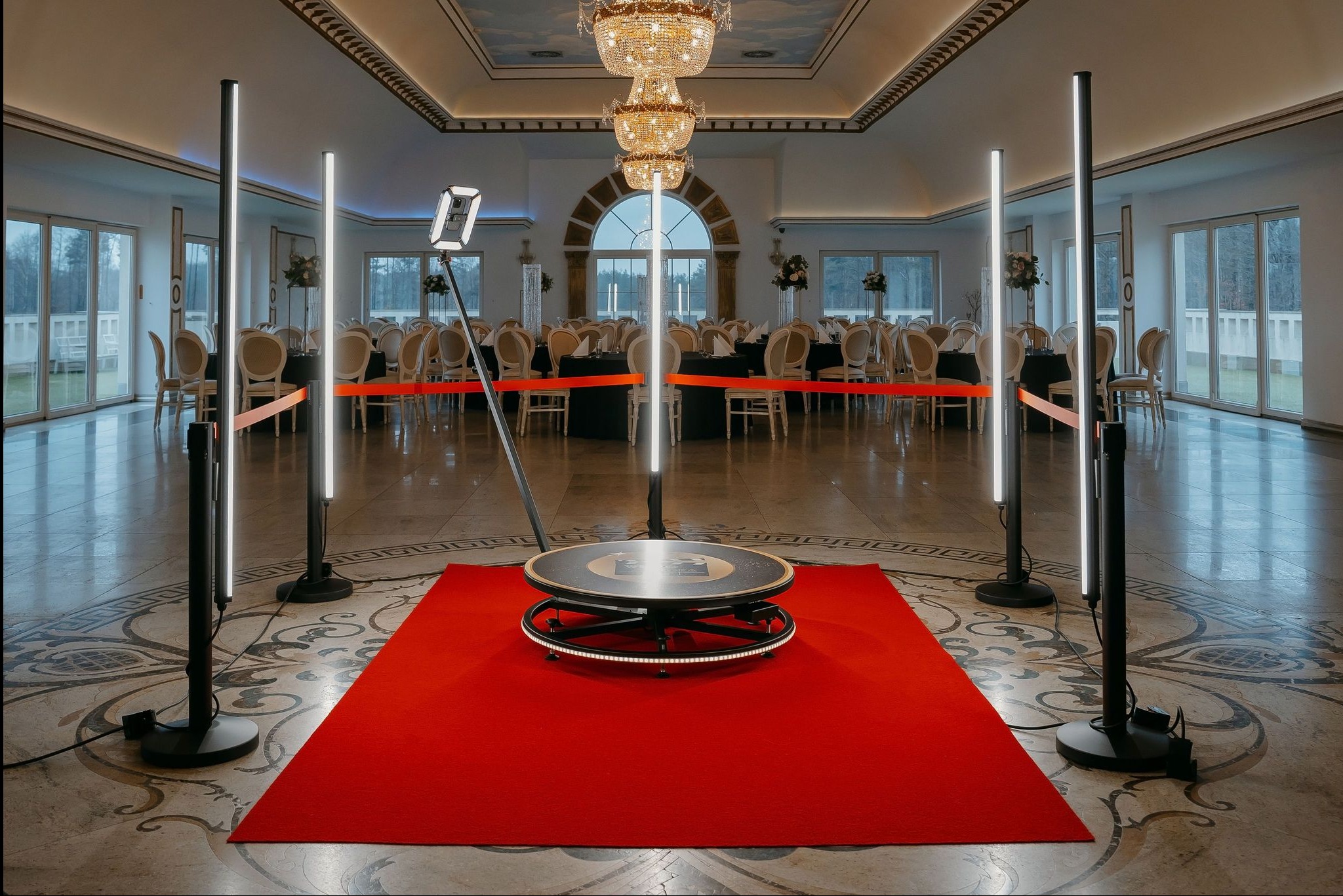 Rotacyjna fotobudka 360 stopni z oświetleniem LED, ustawiona na czerwonym dywanie w eleganckiej sali balowej z kryształowym żyrandolem i dekoracjami stołów.