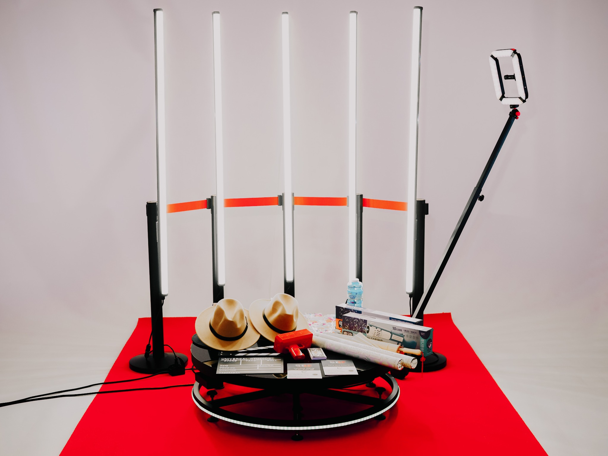 Kompletny zestaw do fotobudki: oświetlenie LED na statywach z czerwonymi taśmami, obrotowa platforma z rekwizytami (kapelusze, klaps filmowy, zabawki) i uchwyt na telefon z dodatkowym oświetleniem,...