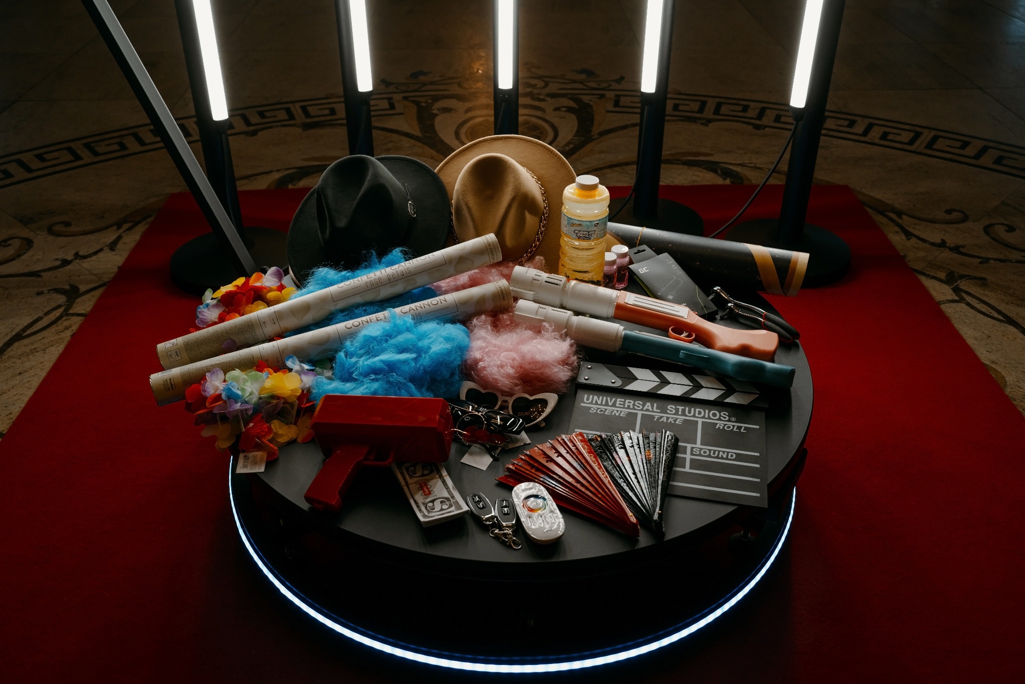 Zestaw rekwizytów do fotobudki: kapelusze, pistolety na konfetti, okulary w kształcie serc, klaps filmowy Universal Studios, wachlarze i hawajskie naszyjniki, ułożone na okrągłym, podświetlanym...