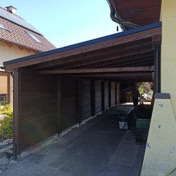 MWJ Adrian Petla - Drewniana konstrukcja zadaszenia przylegająca do budynku, z widocznymi elementami konstrukcyjnymi oraz fragmentem posesji z domem i zielenią. W tle widoczny mężczyzna.
