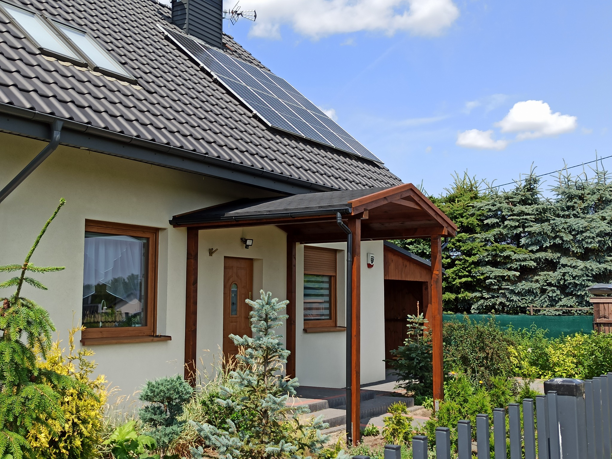 Dom jednorodzinny z drewnianą wiatą nad wejściem, panelami słonecznymi na dachu i zadbanym ogrodem z iglakami.