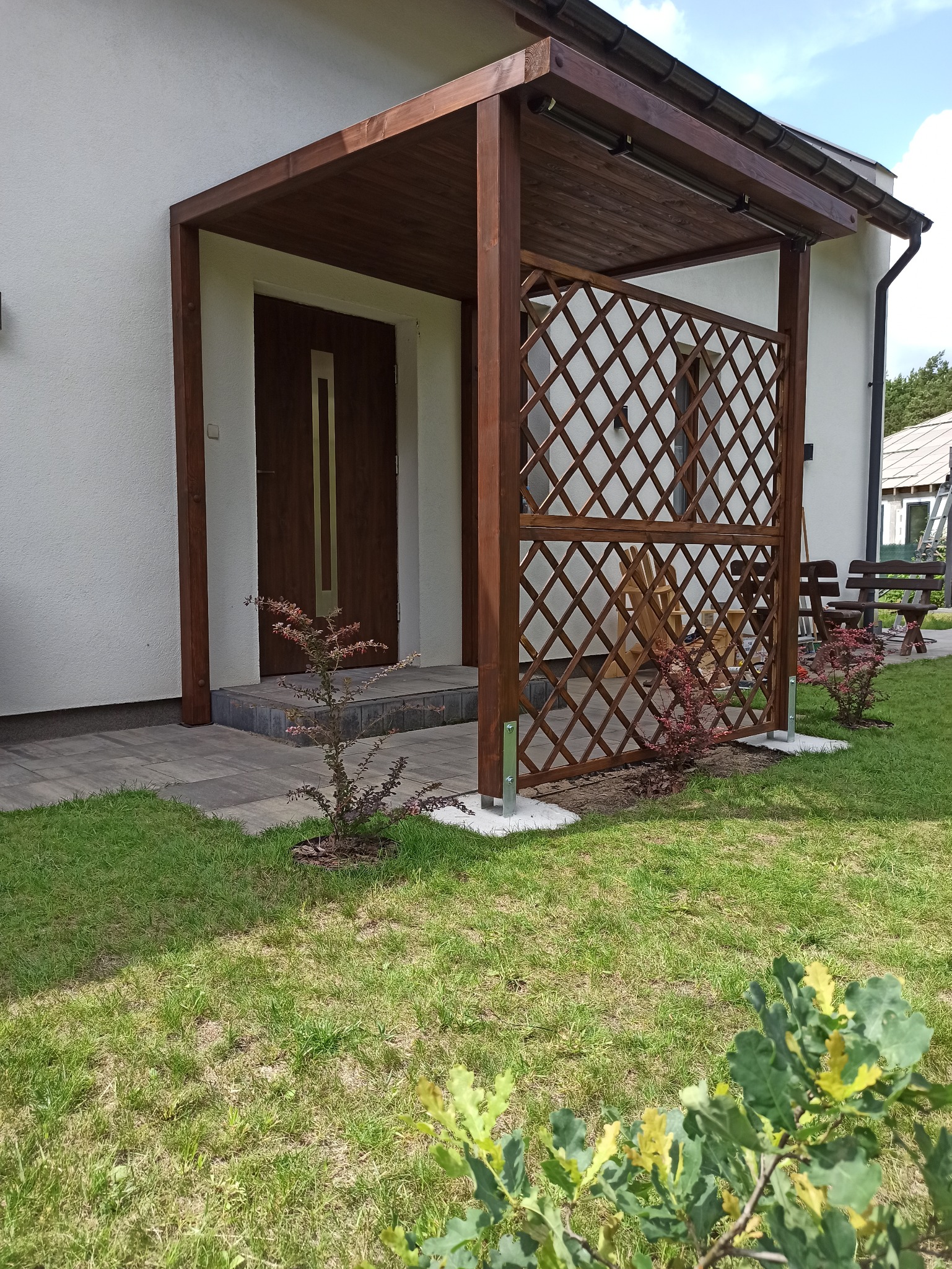 Drewniana konstrukcja z ażurową ścianką przy wejściu do domu, z widocznymi elementami oświetlenia i roślinami ozdobnymi.