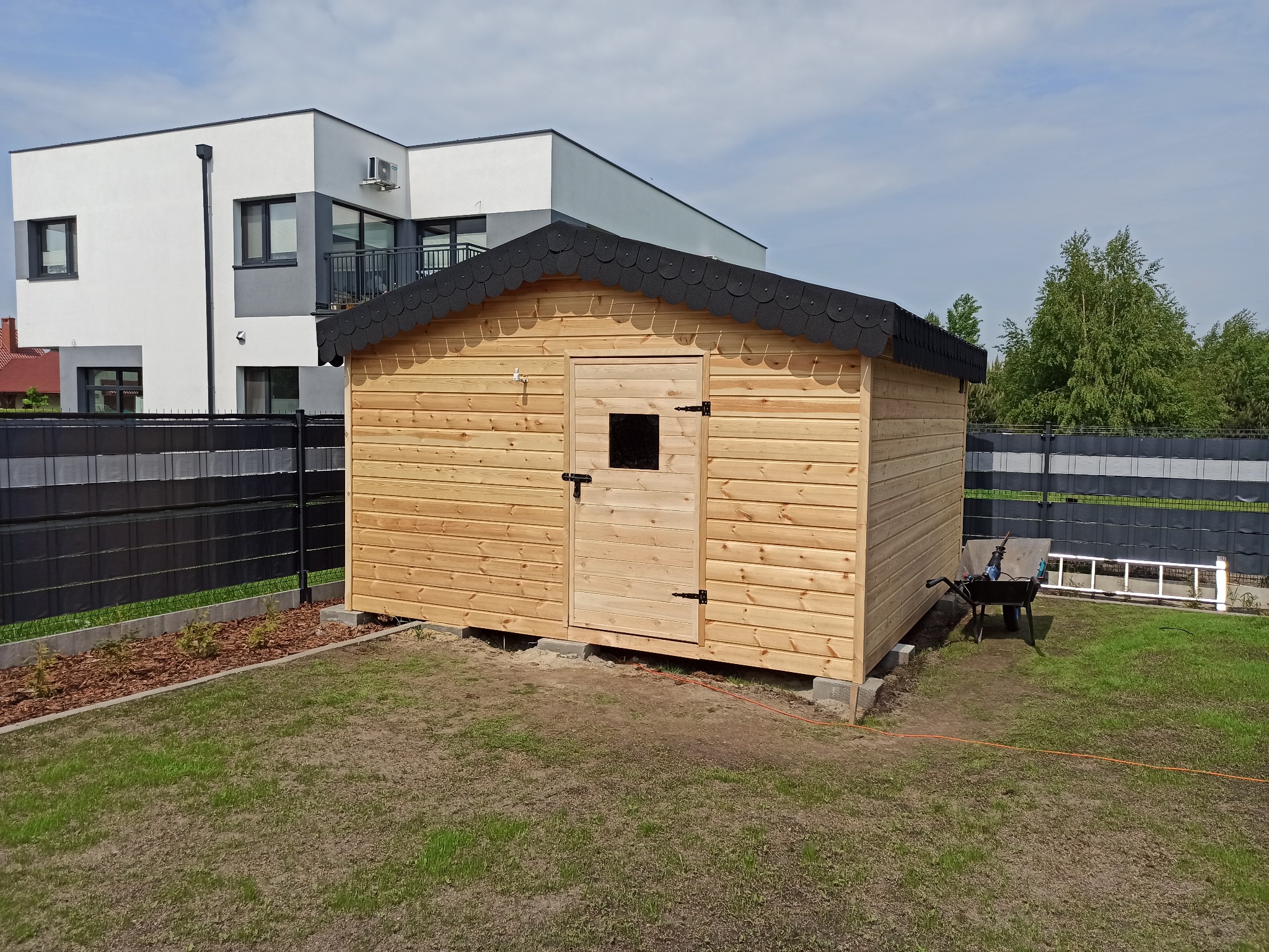 Drewniana altana ogrodowa z czarnym dachem i małym okienkiem w drzwiach, na tle nowoczesnego domu i czarnego ogrodzenia panelowego.
