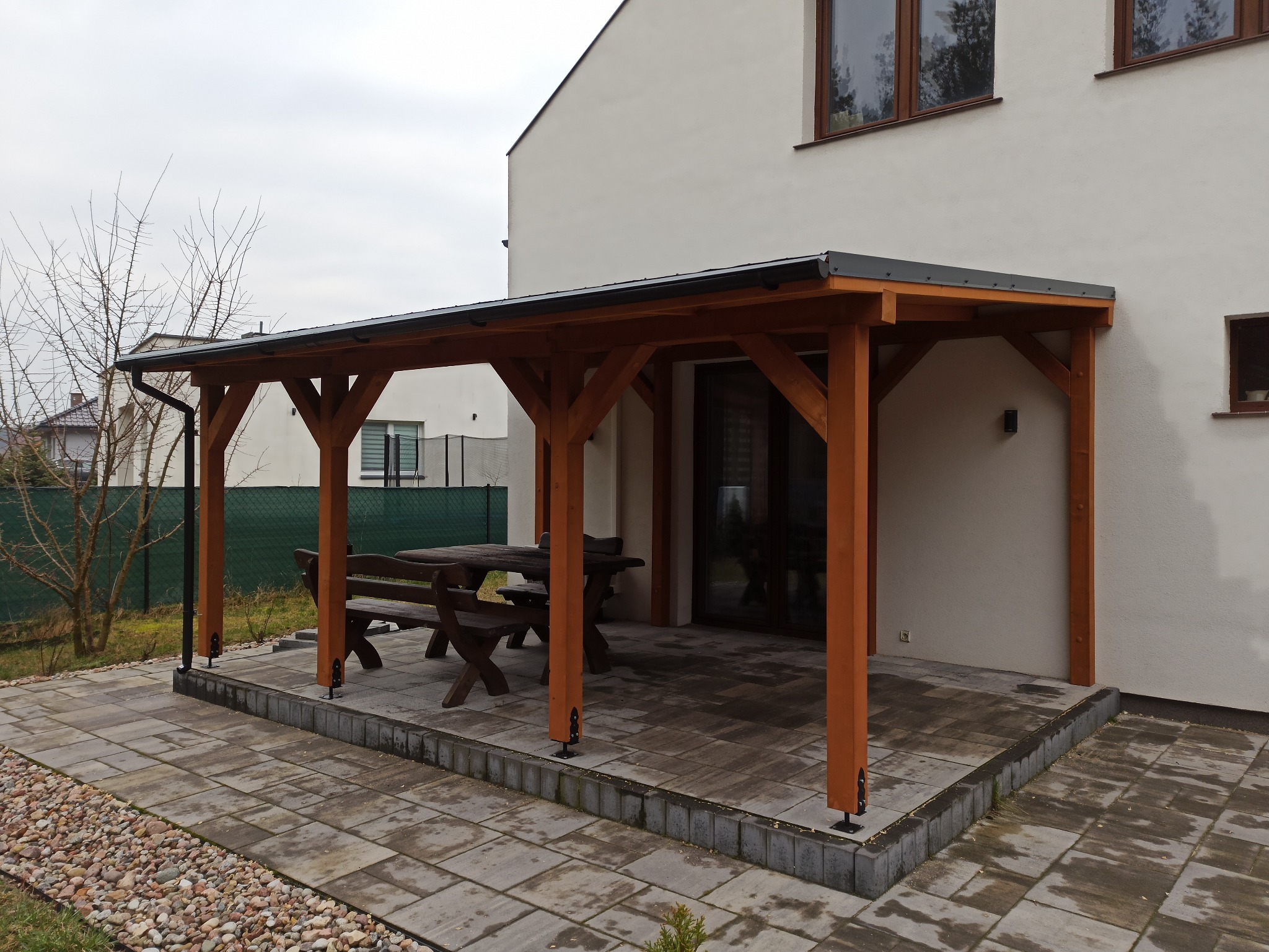 Drewniana pergola z ciemnym dachem przylegająca do białego budynku, z drewnianym stołem i ławkami na wybrukowanym tarasie.