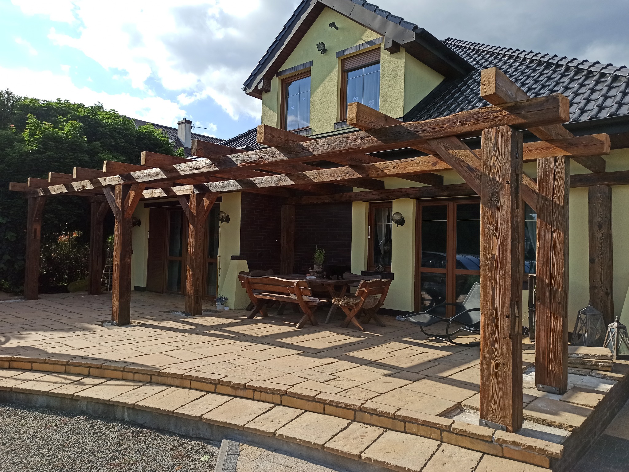 Drewniana pergola z masywnych belek nad tarasem z kamienną posadzką, widok na dom z oknami i zielenią w tle, meble ogrodowe pod pergolą.
