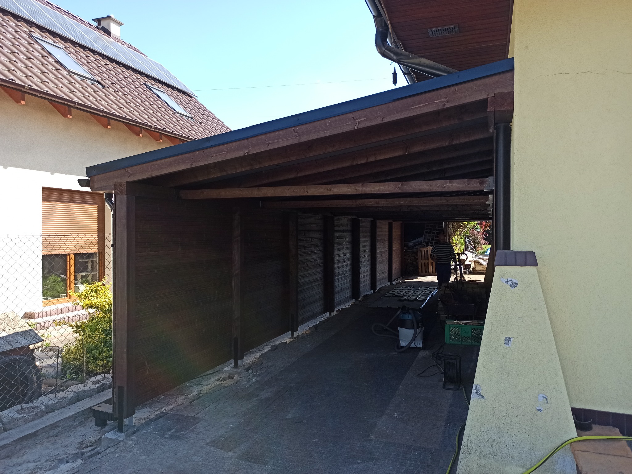 Drewniana konstrukcja zadaszenia przylegająca do budynku, z widocznymi elementami konstrukcyjnymi oraz fragmentem posesji z domem i zielenią. W tle widoczny mężczyzna.
