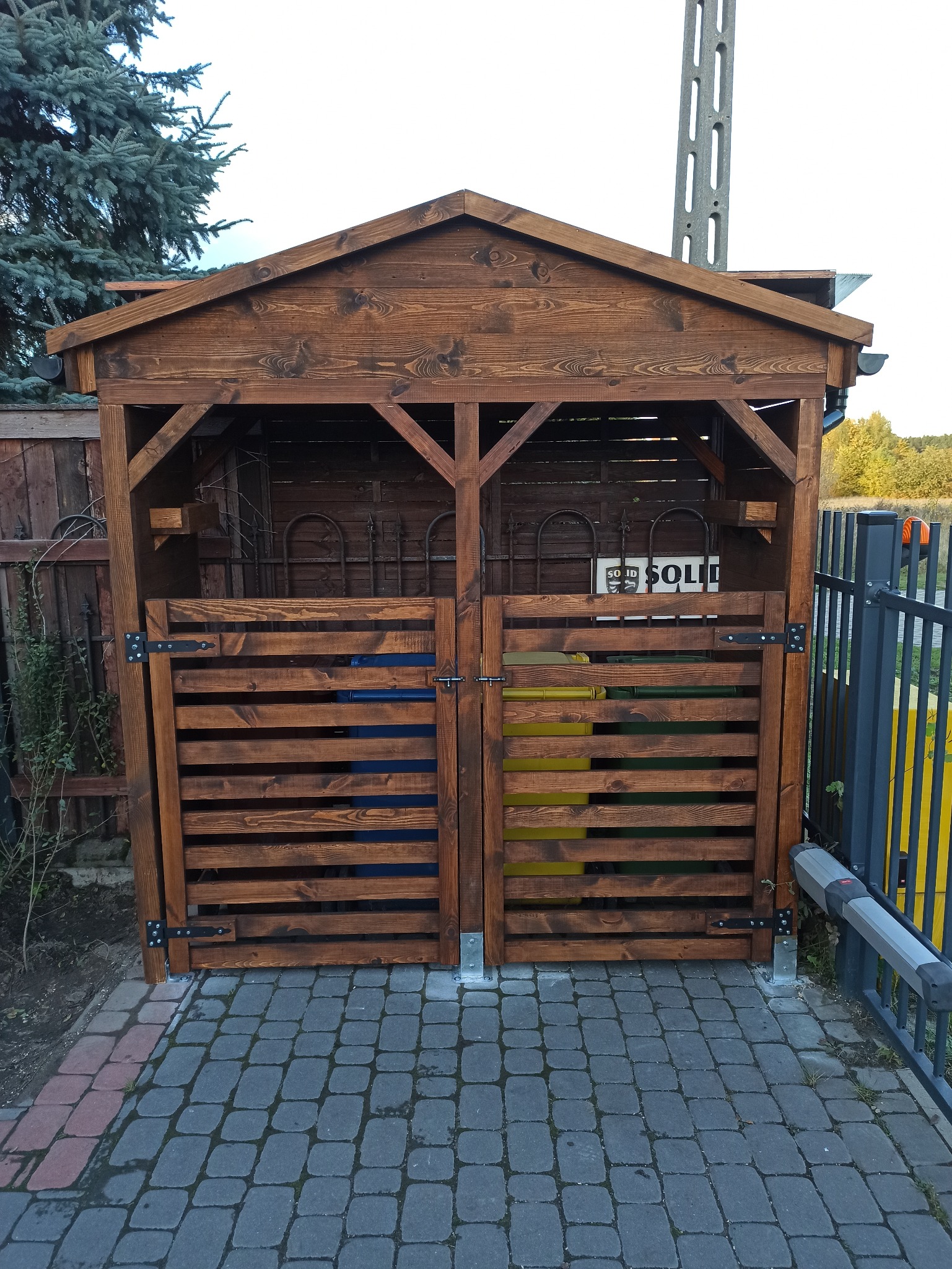 Drewniana zabudowa na pojemniki do segregacji śmieci z zadaszeniem, z widocznymi pojemnikami na szkło i plastik, na tle kostki brukowej i ogrodzenia.