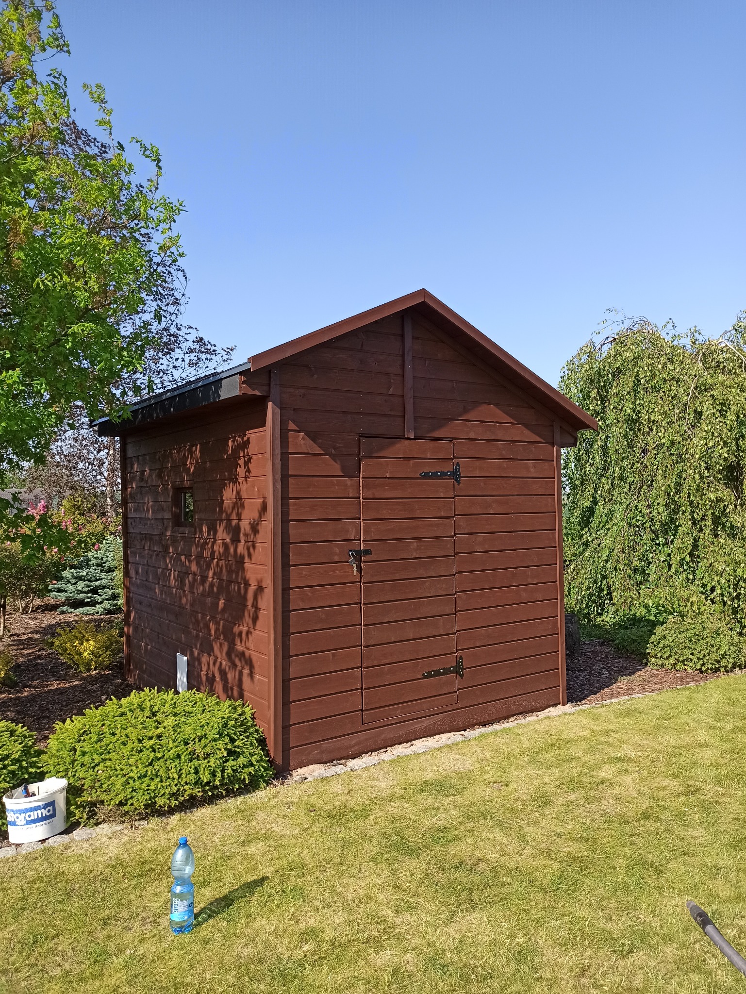 Brązowy, drewniany domek narzędziowy z dwuspadowym dachem na zadbanym trawniku, otoczony zielenią, z widocznym wiadrem z farbą i butelką wody.