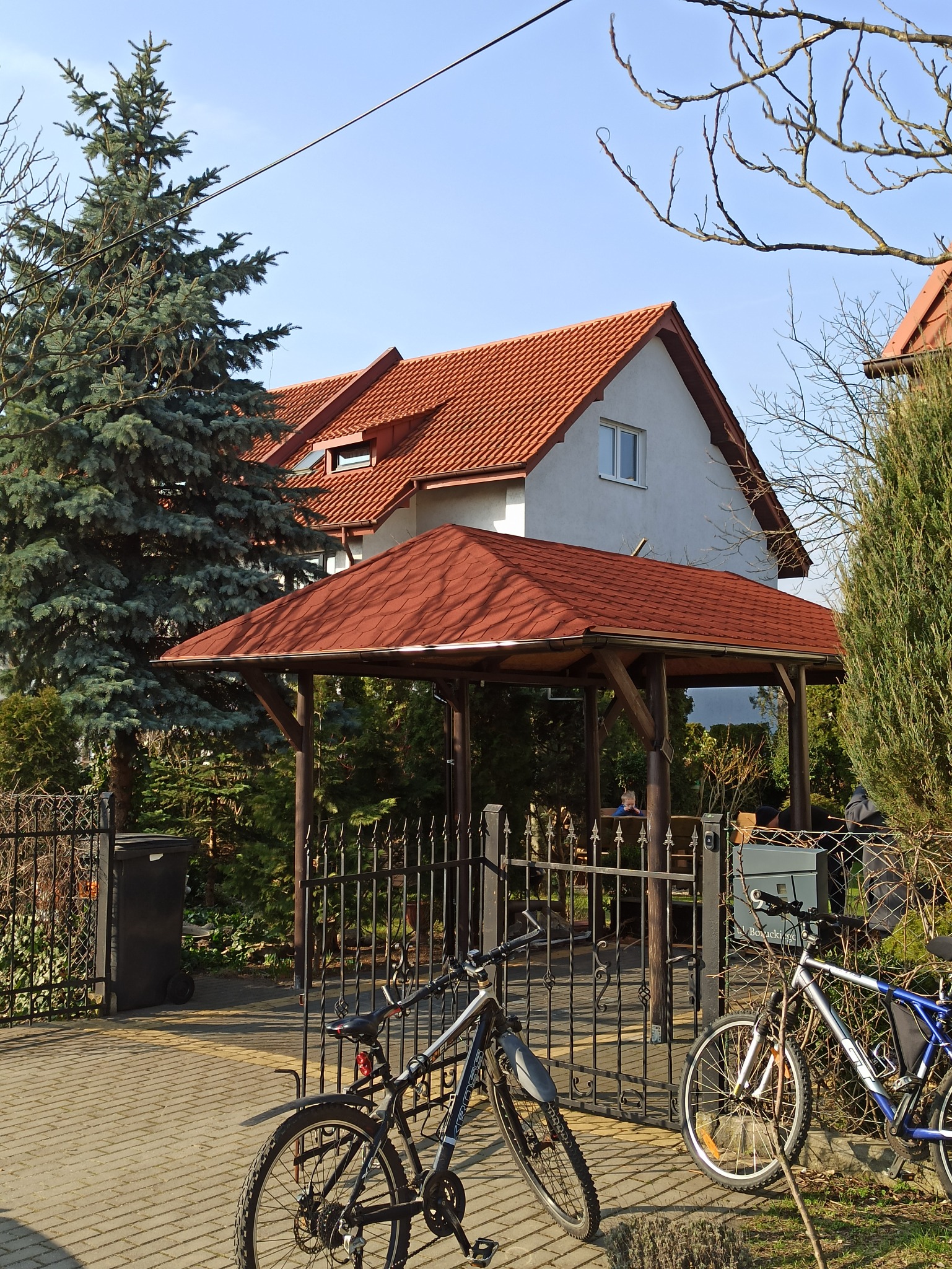 Drewniana konstrukcja z czerwoną dachówką w ogrodzie, widoczna przez metalową bramę, z rowerami opartymi o ogrodzenie.