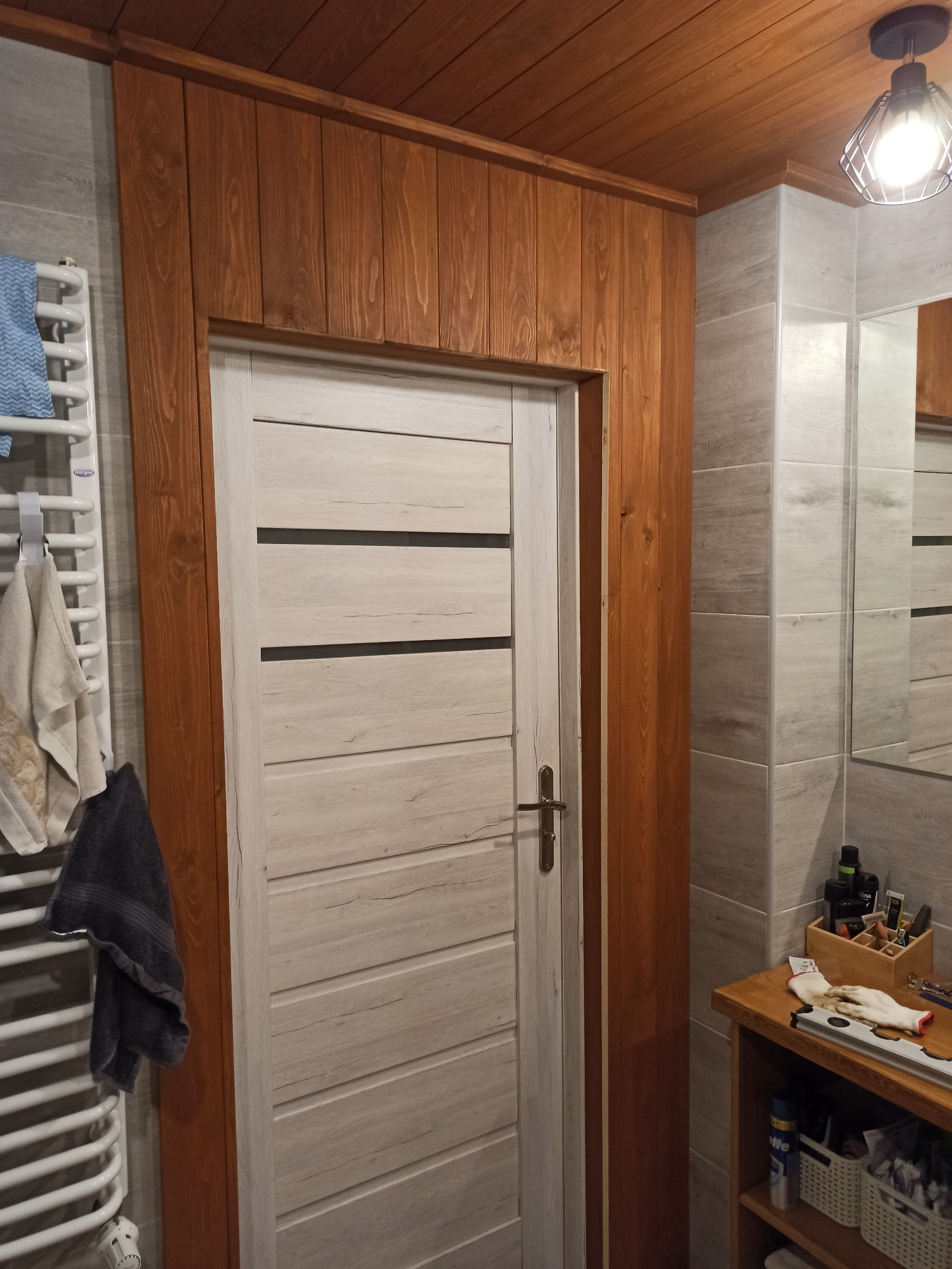 Wykończona łazienka z drewnianą boazerią, sufitem i obramowaniem drzwi w kolorze miodowego dębu, jasnoszare płytki imitujące drewno na ścianach, białe drzwi z poziomymi czarnymi paskami i grzejnik...