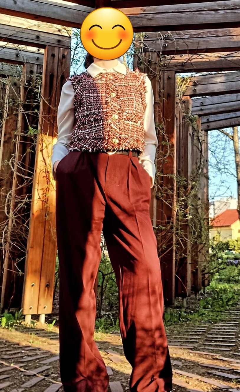 Osoba w bordowych spodniach i tweedowej kamizelce na tle drewnianej konstrukcji z pnączami. Stylizacja w plenerze, naturalne światło.