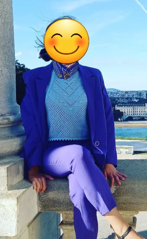 Kobieta w kobaltowym garniturze i błękitnym swetrze pozuje na tle nadmorskiego krajobrazu. Uwagę zwraca stylizacja kolorystyczna i dopasowanie stroju do otoczenia.