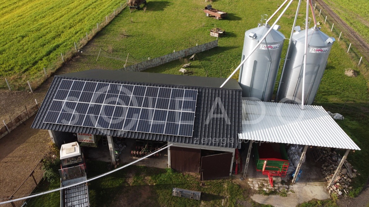 Widok z góry na budynek gospodarczy z panelami fotowoltaicznymi na dachu, obok silosy zbożowe, traktor i maszyny rolnicze na terenie gospodarstwa.