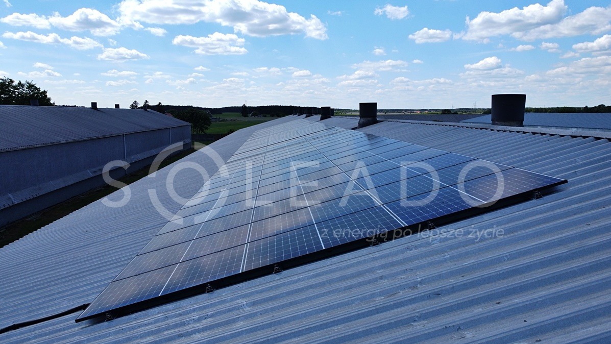 Instalacja fotowoltaiczna na dachu budynku gospodarczego, widok z góry, niebieskie panele słoneczne częściowo odbijają niebo, w tle pola i lasy.