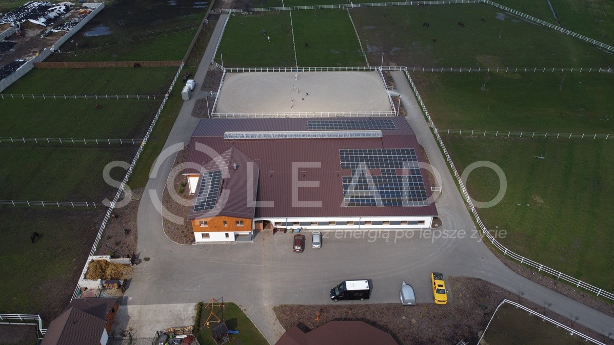 Widok z lotu ptaka na budynek stajni z panelami słonecznymi na dachu, przyległy plac treningowy dla koni i pastwiska otoczone białym ogrodzeniem.
