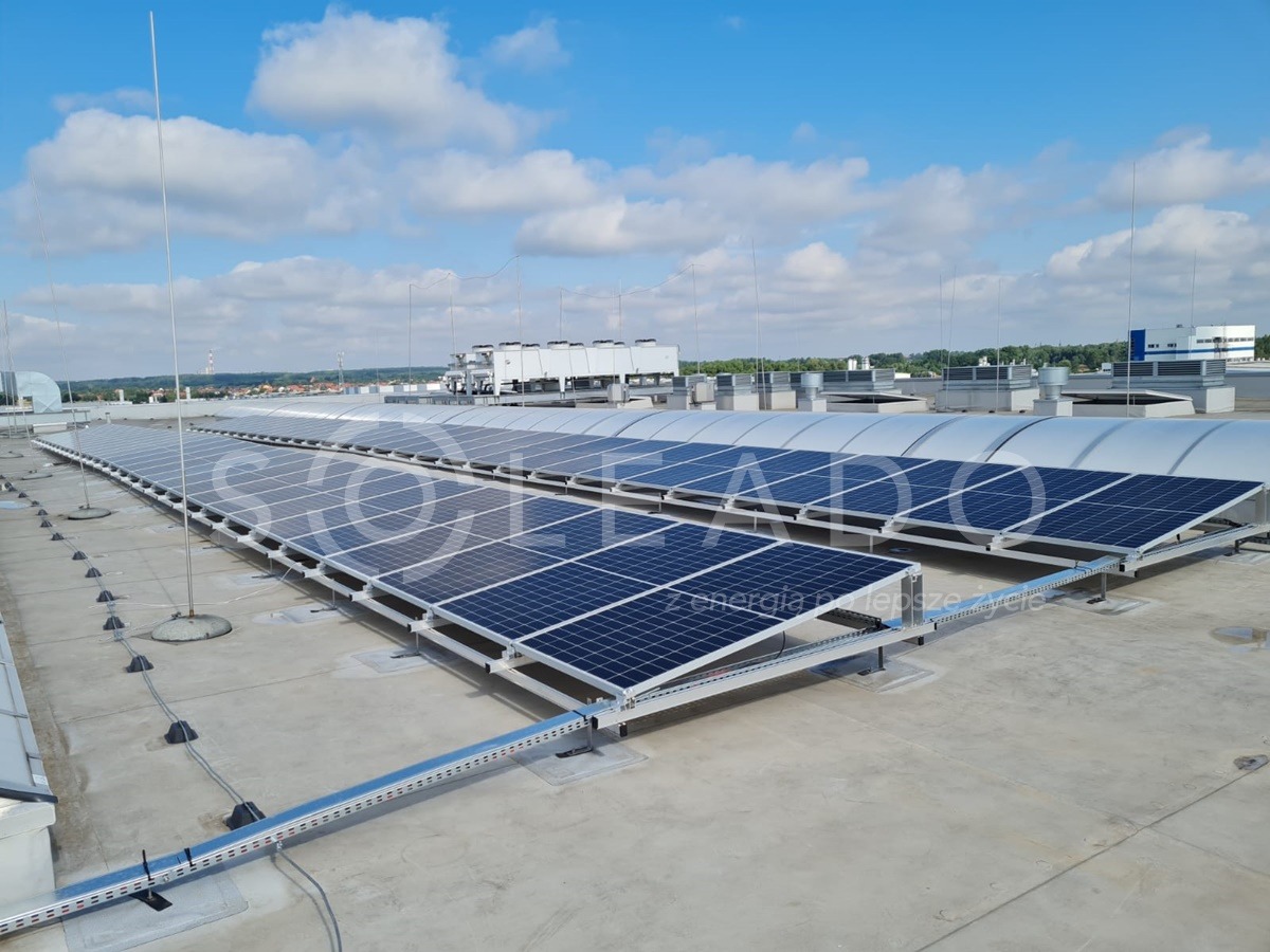 Rozległa instalacja paneli słonecznych na płaskim dachu budynku przemysłowego, widoczne zabezpieczenia odgromowe i systemy wentylacyjne w tle, błękitne niebo z obłokami.