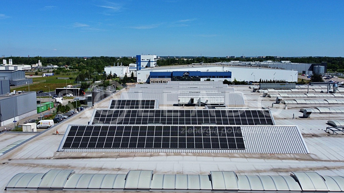 Widok z góry na panele słoneczne zamontowane na dachu dużego budynku przemysłowego, w tle zabudowania fabryczne i zielone tereny.