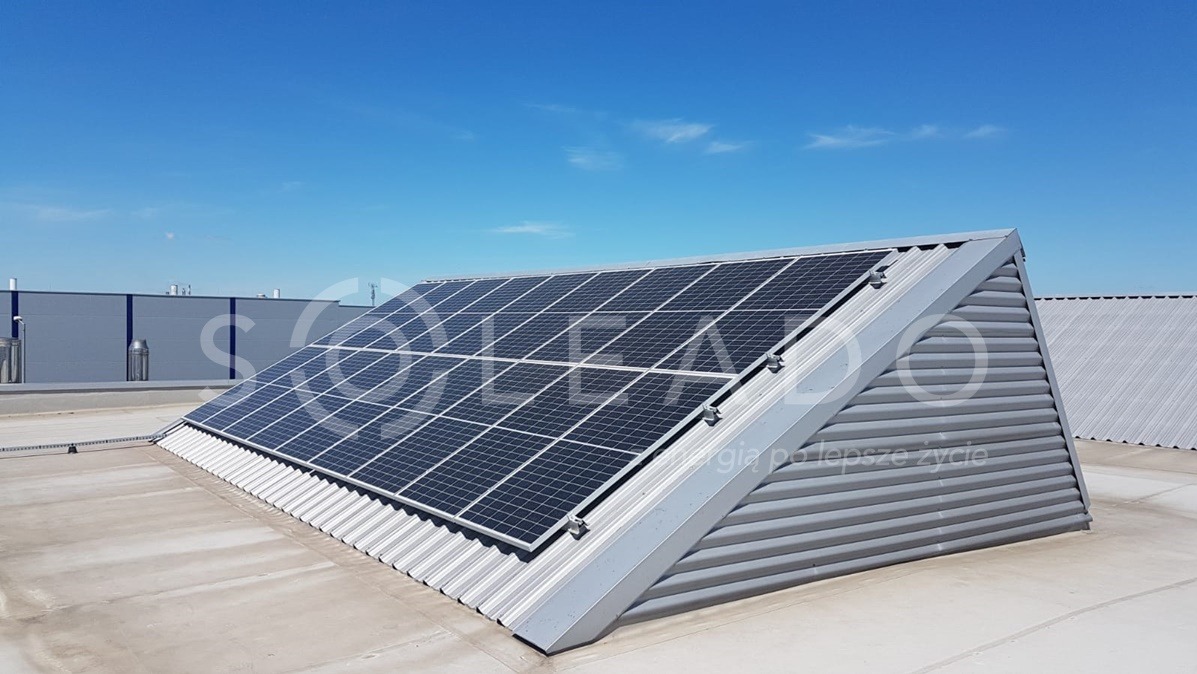 Panele słoneczne zamontowane na skośnym dachu z blachy falistej, na tle błękitnego nieba i budynku przemysłowego.