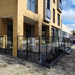 Szklana balustrada z czarnymi słupkami otacza podwyższony taras przy budynku o elewacji z paneli imitujących drewno. Widoczny chodnik z kostki brukowej, fragmenty rabat z ziemią, okna budynku...