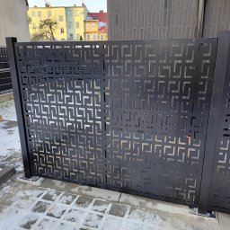 Bos-spaw Tadeusz Sieradzki - Nowoczesne, metalowe ogrodzenie panelowe w kolorze antracytowym z geometrycznym wzorem, częściowo pokryte śniegiem, na tle budynku mieszkalnego.