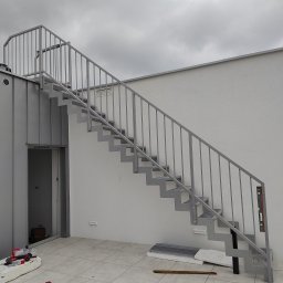 Bos-spaw Tadeusz Sieradzki - Zewnętrzne schody metalowe z szarą balustradą na tle białej ściany i pochmurnego nieba, widoczne narzędzia i materiały budowlane sugerują trwający montaż.