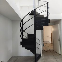 Bos-spaw Tadeusz Sieradzki - Nowoczesne, czarne schody spiralne z metalową balustradą w minimalistycznym wnętrzu z surowym betonem na podłodze i białymi ścianami. Słup schodów owinięty folią ochronną.