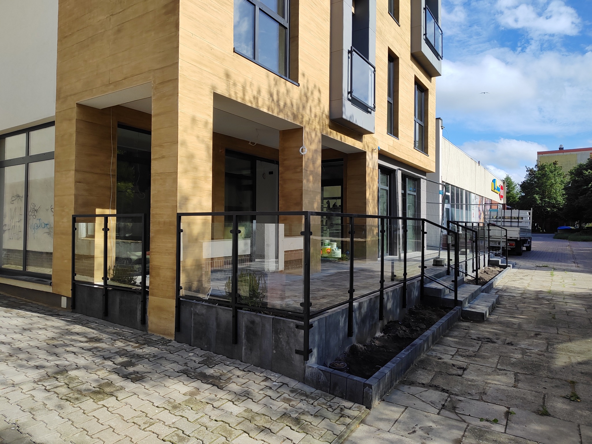 Szklana balustrada z czarnymi słupkami otacza podwyższony taras przy budynku o elewacji z paneli imitujących drewno. Widoczny chodnik z kostki brukowej, fragmenty rabat z ziemią, okna budynku...