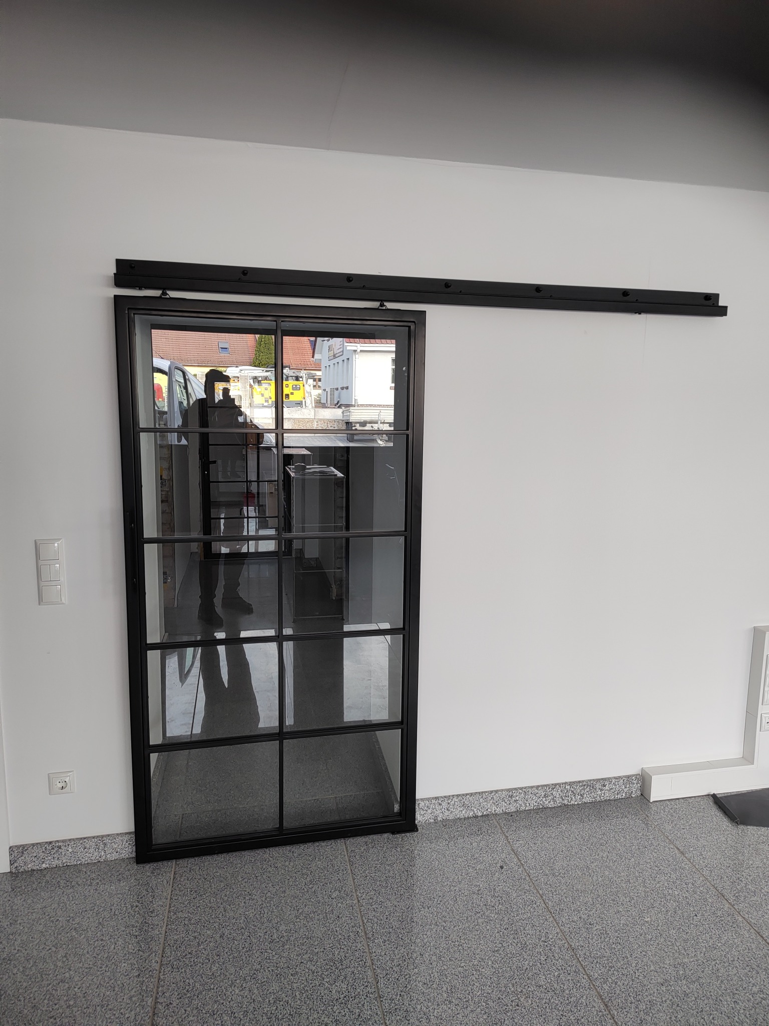 Przesuwne, szklane drzwi w czarnej ramie z podziałami, zamontowane na szynie przy białej ścianie. W szkle widoczne odbicie otoczenia zewnętrznego i wnętrza pomieszczenia.