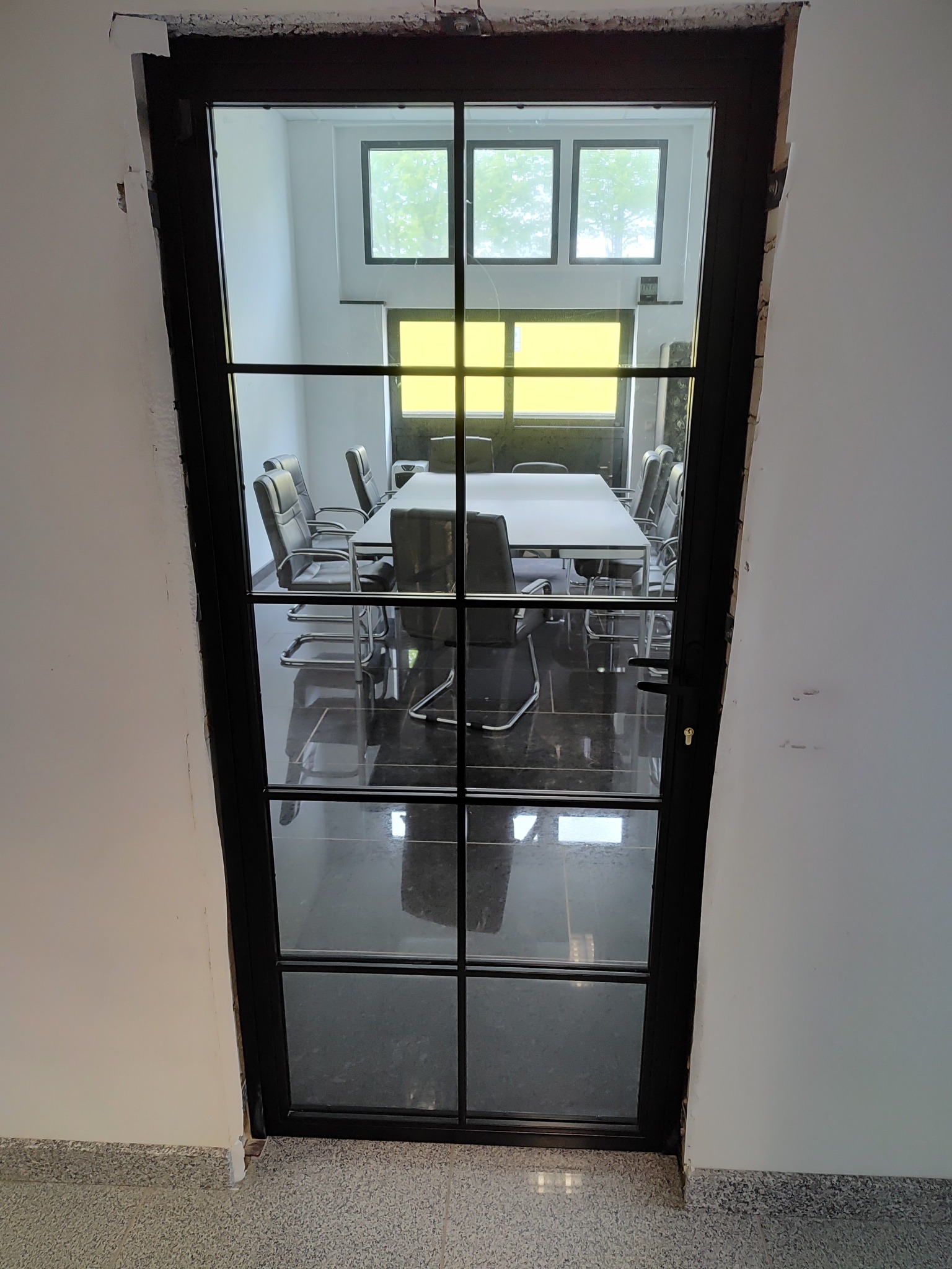 Czarne, przeszklone drzwi wewnętrzne z podziałami szprosami, widok na salę konferencyjną z białym stołem i krzesłami, surowy montaż w białej ścianie.