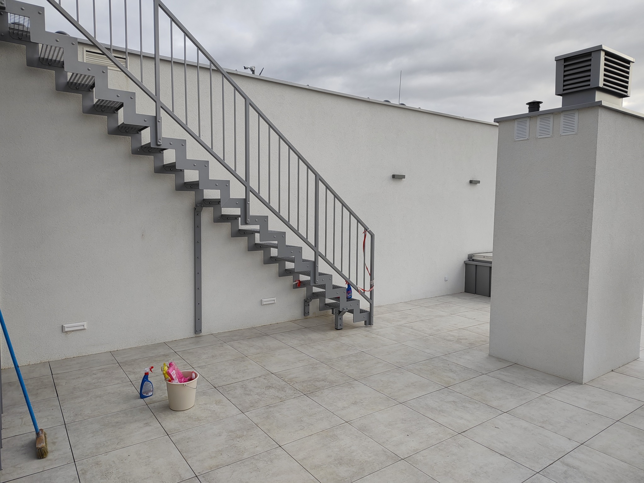 Zewnętrzne metalowe schody z szarą balustradą prowadzące na dach, widok z dołu, na dachu wiadro ze środkami czystości.