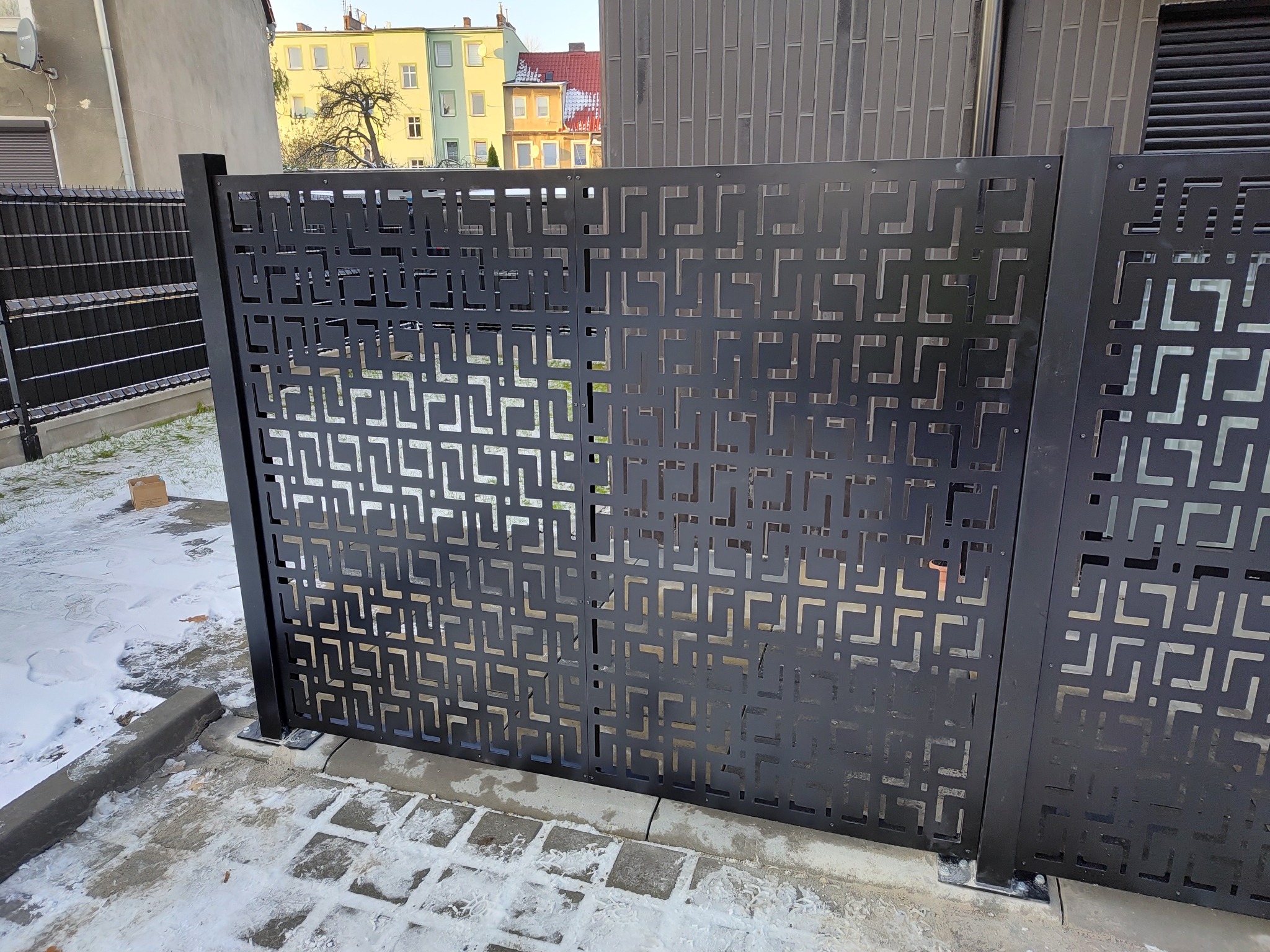 Nowoczesne, metalowe ogrodzenie panelowe w kolorze antracytowym z geometrycznym wzorem, częściowo pokryte śniegiem, na tle budynku mieszkalnego.