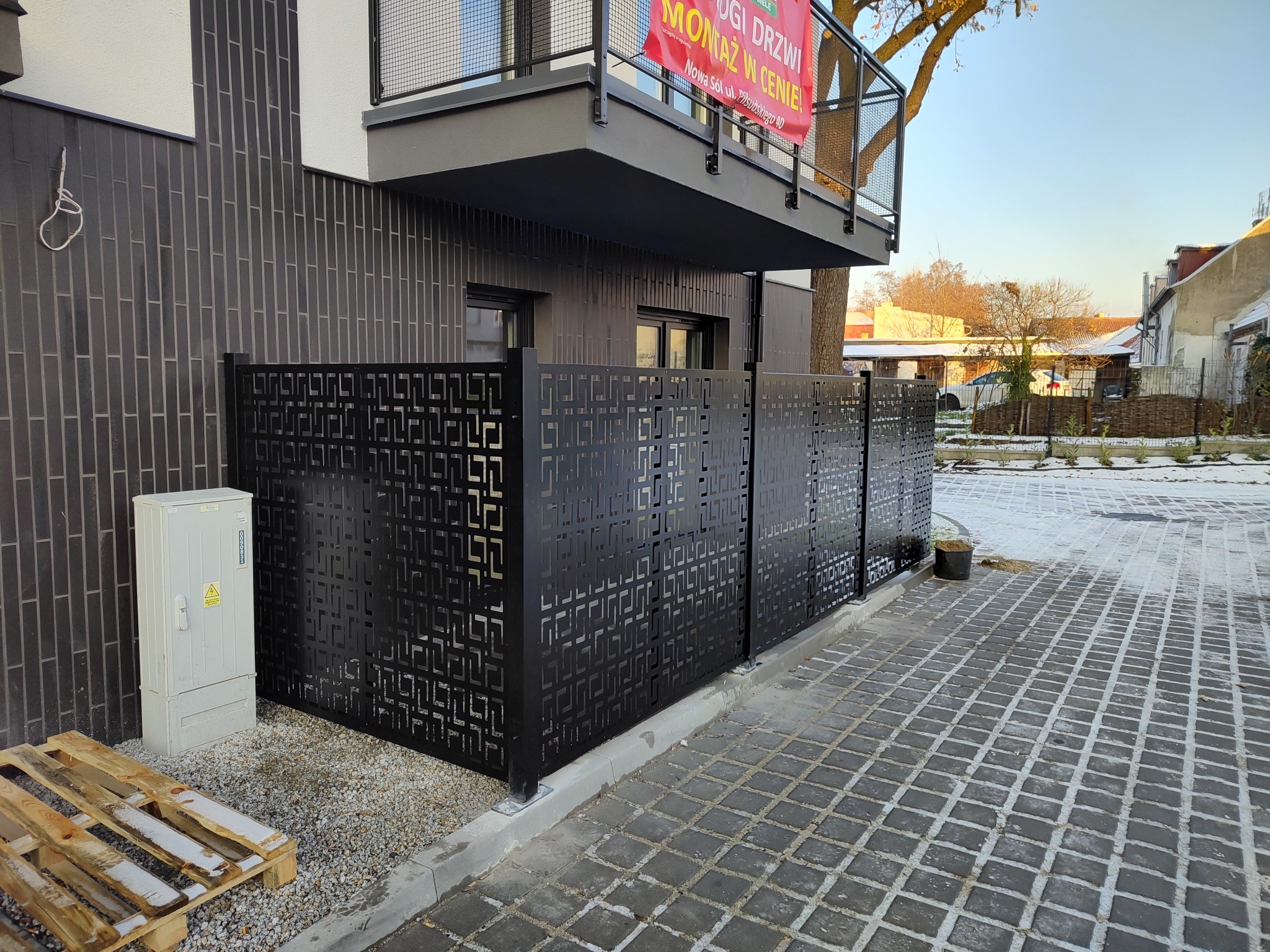 Nowoczesne, czarne ogrodzenie panelowe z geometrycznym wzorem, oddzielające posesję od chodnika z kostki brukowej, z widoczną skrzynką elektryczną i paletą w tle, w zimowej scenerii.