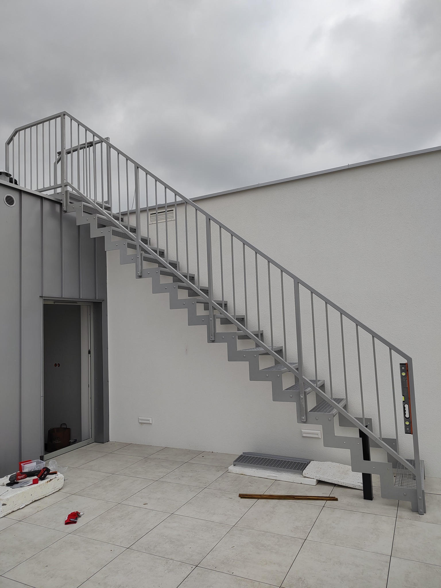 Zewnętrzne schody metalowe z szarą balustradą na tle białej ściany i pochmurnego nieba, widoczne narzędzia i materiały budowlane sugerują trwający montaż.