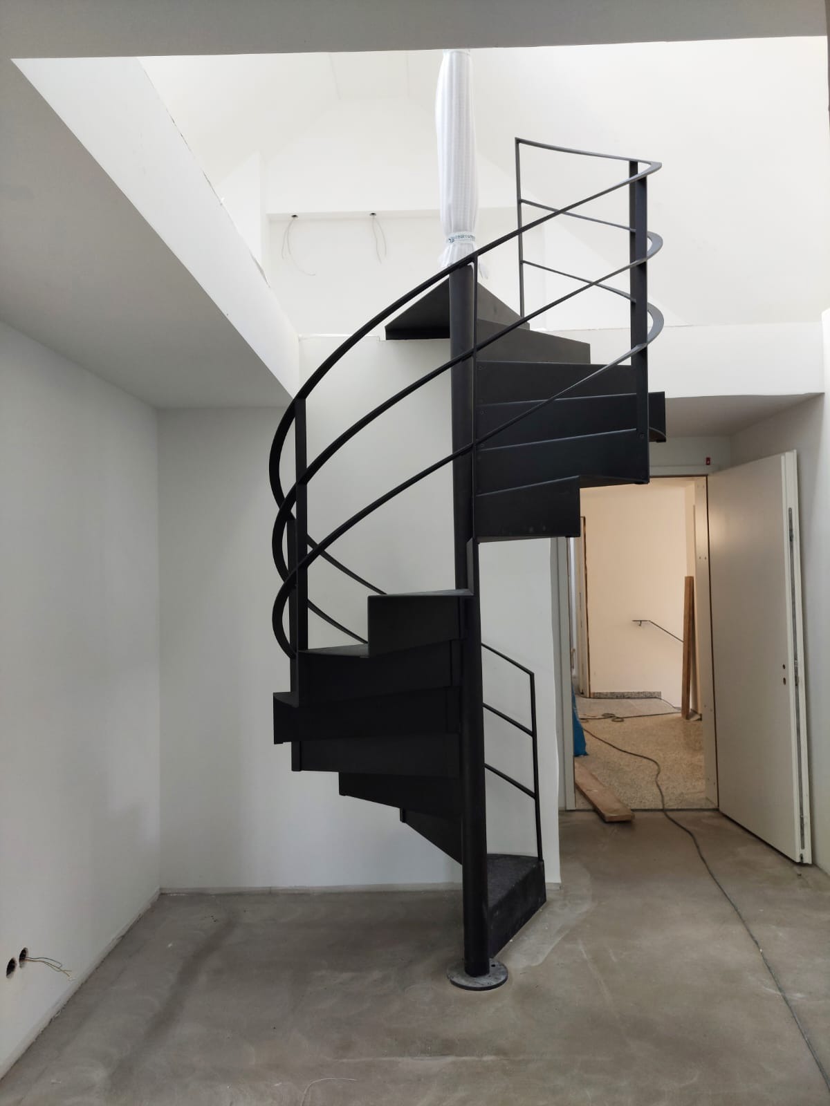 Nowoczesne, czarne schody spiralne z metalową balustradą w minimalistycznym wnętrzu z surowym betonem na podłodze i białymi ścianami. Słup schodów owinięty folią ochronną.