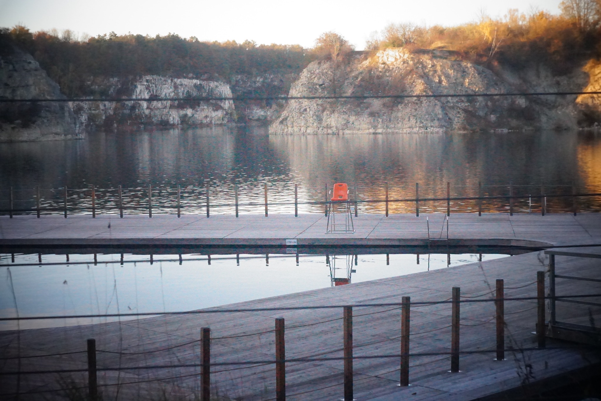 Spokojny krajobraz: Pomost nad wodą z krzesłem ratownika na tle skalistego brzegu i odbicia w wodzie.