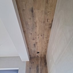 Bartłomiej Maczuga - Ściana i sufit wyłożone płytkami imitującymi drewno, z widocznymi otworami na oświetlenie i wystającymi kablami, oraz fragment ściany z szarymi płytkami i oknem w tle.