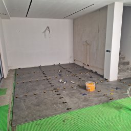 Bartłomiej Maczuga - Układanie szarych płytek podłogowych z użyciem systemu poziomowania, widoczny pojemnik z klinami, podkład z zielonej pianki i surowe ściany w trakcie remontu.