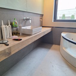 Bartłomiej Maczuga - Łazienka w trakcie wykańczania: umywalka nablatowa, silikony, narzędzia i płytki imitujące drewno na blacie oraz szare płytki na ścianach i podłodze, widoczna wanna z hydromasażem i okno z widokiem...